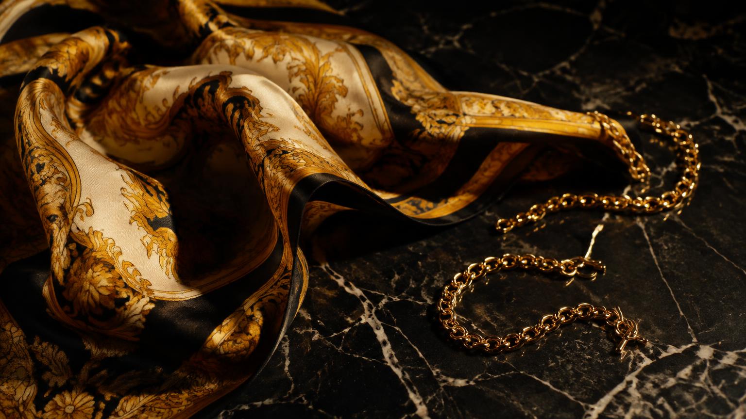 Silk scarf with gold-toned baroque print and gold chain link necklace on black marble.